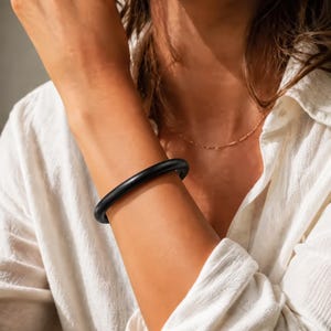 Pode incluir: Uma pulseira rígida preta, sólida e lisa, com um perfil arredondado. A pulseira apresenta um design circular contínuo com um acabamento mate, usada no pulso. É uma peça de joalharia simples e sem adornos, com uma espessura constante.