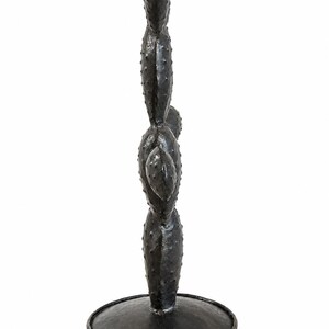 May include: A dark charcoal gray metal sculpture shaped like a prickly pear cactus with multiple stacked, oval-shaped pads featuring small raised bumps across the surface. The cactus stands upright on a circular, flat metal base of the same color.
