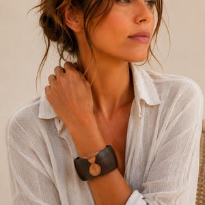 May include: A wide cuff bracelet featuring a dark brown, smooth exterior with a contrasting circular light brown wood inlay centered on the front. The bracelet has a rigid, open-ended design that wraps around the wrist, showcasing a natural wood grain texture.
