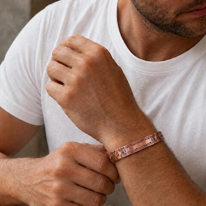 Può includere: Un bracciale a fascia in rame caratterizzato da una finitura martellata e testurizzata, con dettagli ossidati scuri lungo i bordi e un motivo geometrico centrale. La fascia metallica presenta una lucentezza polita e un profilo largo e piatto.