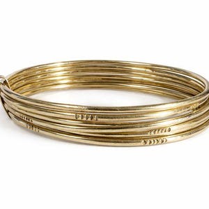 Peut inclure: Un ensemble de bracelets de couleur or. Les bracelets sont fins, ronds et semblent être en métal. Certains bracelets présentent des motifs décoratifs gravés. Les bracelets sont empilés sur un fond blanc.