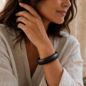 May include: A thick, solid black bangle bracelet with a smooth, rounded surface. The bracelet features a circular shape and a matte finish, encircling the wrist. The design is simple and uniform, showcasing a dark, deep black color throughout.