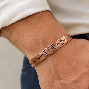 Peut inclure: Un bracelet manchette en cuivre orné d'une série de motifs circulaires en relief et de lignes gravées. Le métal présente une teinte brun-rouge chaleureuse avec des zones en creux assombries soulignant les détails géométriques complexes.