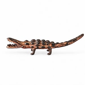 May include: A small, metallic alligator figurine with a textured, dark bronze-toned finish. The alligator is depicted in a horizontal standing position with its mouth open, revealing sharp teeth, and a bumpy, scaled back surface.