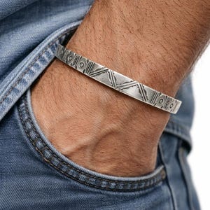 Peut inclure: Un bracelet manchette argenté avec des motifs géométriques. Le bracelet est porté au poignet, avec une poche en jean en arrière-plan. Le bracelet présente des lignes gravées et des motifs circulaires.