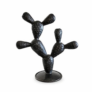 May include: A black, textured cactus sculpture featuring multiple rounded, paddle-shaped segments with small, raised bumps across the surface. The cactus is mounted on a circular, flat base, creating a stable, upright, and decorative object.