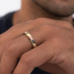 May include: A ring featuring a multi-toned band with alternating diagonal segments of polished gold, rose gold, and a dark, textured metallic finish. The band has a smooth, rounded profile and a consistent width, showcasing a blend of metallic colors.