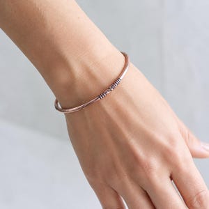 Peut inclure: Un bracelet jonc fin de couleur cuivre, doté d'une finition lisse et polie, comportant une petite section décorative texturée avec des rainures sombres et creusées qui créent un contraste subtil avec la surface métallique du bracelet.