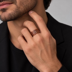 May include: A multi-band ring crafted from polished copper, featuring several thin, stacked circular wires that create a layered appearance. The ring has a warm, metallic reddish-brown hue and a smooth, uniform finish across all the individual bands.