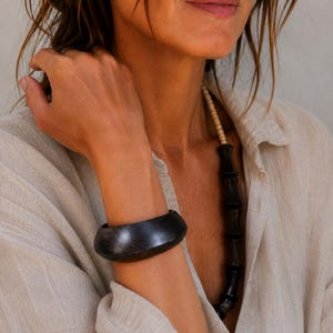 May include: A wide, smooth, dark brown wooden bangle bracelet with a curved, rounded profile. The bracelet features a visible wood grain texture and a polished finish, worn on the wrist alongside a necklace with dark wooden beads.