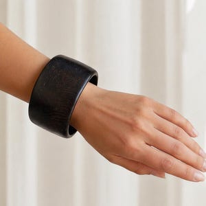May include: A wide, black wooden bracelet is worn on a wrist. The bracelet is a solid, dark color with a smooth, rounded shape. The wood grain is visible. The bracelet is approximately 2.5 inches wide.