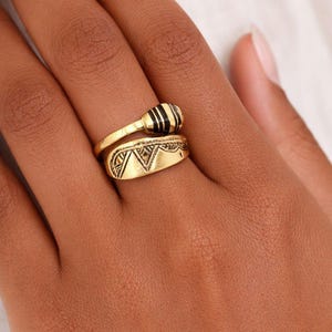 Può includere: Un anello aperto color oro caratterizzato da un'estremità arrotondata e liscia con strisce orizzontali nere e un'estremità più ampia e piatta incisa con una serie di motivi triangolari e dettagli lineari lungo il bordo superiore.