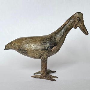 May include: A bronze bird sculpture with a long neck and beak, standing on two feet. The bird has a textured surface with shades of brown and grey, suggesting age and detail. The sculpture is a decorative item.