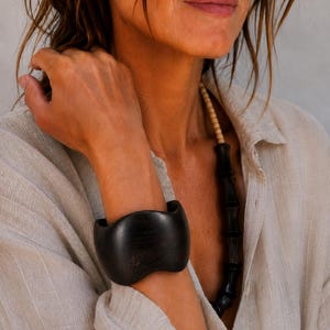 May include: A wide, dark brown wooden cuff bracelet with a smooth, undulating wave-like silhouette. The bracelet features a visible natural wood grain pattern and is worn alongside a necklace composed of small, cylindrical dark wood beads.
