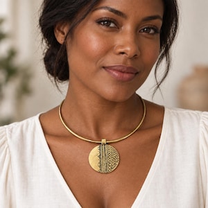Op de afbeelding: Een goudkleurige metalen choker-halsketting met een ronde hanger. De hanger heeft aan één kant een gladde gouden afwerking en aan de andere kant een gestructureerd zwart en goud patroon van verticale lijnen, driehoeken en kleine cirkels.