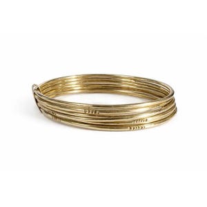 May include: A set of five thin, circular gold-toned metal bangles stacked together. Each bangle features a smooth, polished surface with a small, repeating pattern of etched triangular markings along one section of the outer circumference.