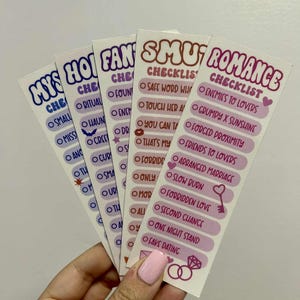 Puede incluir: Una mano sosteniendo cinco pegatinas rectangulares con texto. Las pegatinas tienen un fondo blanco con texto rosa, morado y marrón. Las pegatinas están etiquetadas como "MYS", "HOT", "FAN", "SMUT" y "ROMANCE" con listas de verificación de tropos románticos.