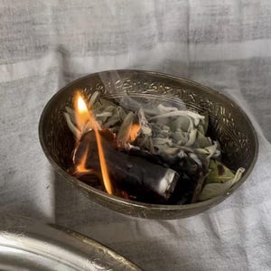 Op de afbeelding: Een kleine, sierlijke zilveren kom met brandende salie en een stuk hout. De vlammen zijn fel oranje en geel, met slierten rook. De kom staat op een wit en grijs gestreepte doek.