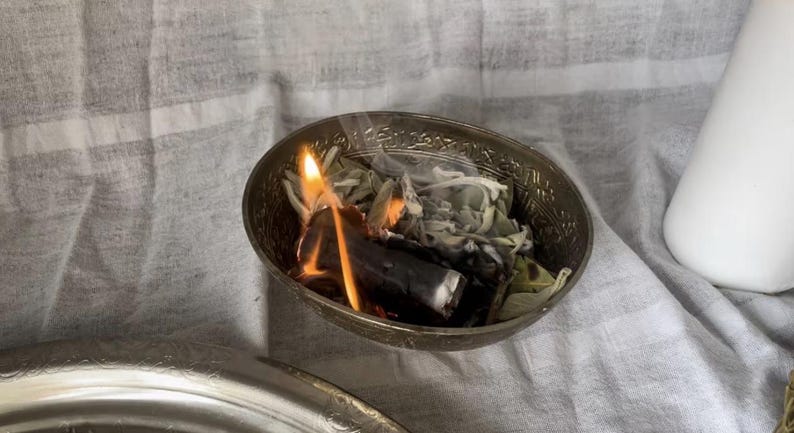 Op de afbeelding: Een kleine, sierlijke zilveren kom met brandende salie en een stuk hout. De vlammen zijn fel oranje en geel, met slierten rook. De kom staat op een wit en grijs gestreepte doek.