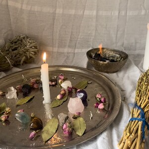 K&ouml;nnte beinhalten: Ein silbernes Tablett mit wei&szlig;en Kerzen, getrockneten Kr&auml;utern und Kristallen, die darauf angeordnet sind. Das Tablett ist von wei&szlig;em Stoff und einem B&uuml;ndel getrockneter Kr&auml;uter umgeben.