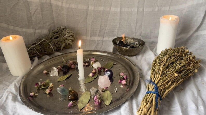 May include: A spiritual arrangement featuring lit candles, a metal tray with crystals and dried flowers, a bundle of sage, and a burning bowl. The scene is set on a white cloth, creating a serene and mystical ambiance.
