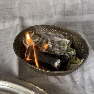 May include: A small, ornate metal bowl filled with burning sage and a piece of charcoal. The flames are bright orange and yellow, with wisps of smoke rising. The bowl is set on a white and gray patterned cloth.