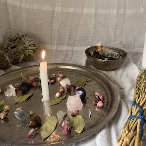 Op de afbeelding: Een stilleven met een brandende witte kaars, een zilveren dienblad met kristallen en gedroogde bloemen, een bundel gedroogde kruiden en een kom met brandende wierook. De sc&egrave;ne is geplaatst op een witte doek.