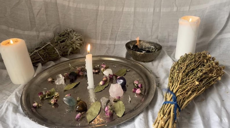 Op de afbeelding: Een stilleven met een brandende witte kaars, een zilveren dienblad met kristallen en gedroogde bloemen, een bundel gedroogde kruiden en een kom met brandende wierook. De sc&egrave;ne is geplaatst op een witte doek.