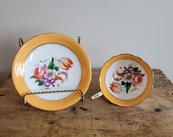 May include: A vintage teacup and saucer set with a yellow rim and floral pattern. The floral pattern features pink, white, and purple flowers with green leaves. The saucer has a scalloped edge.