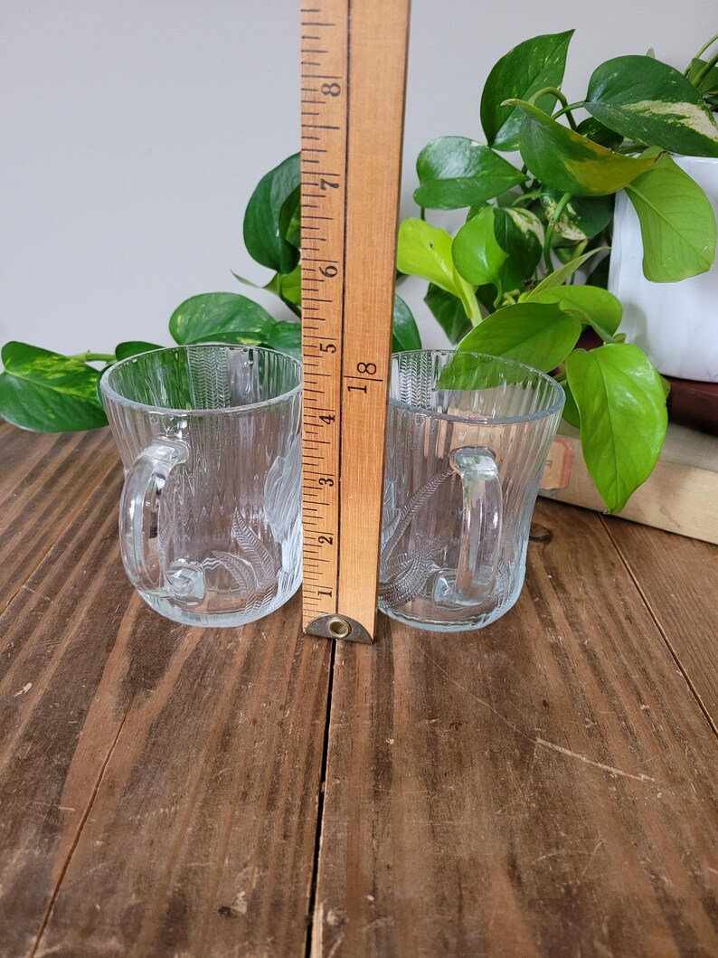 Puede incluir: Dos tazas de vidrio transparente con un dise&ntilde;o texturizado y asas. Las tazas est&aacute;n sobre una superficie de madera.