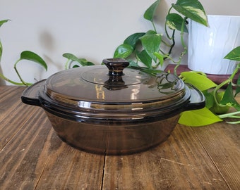 May include: A brown glass casserole dish with a lid. The dish has a square knob on the lid and is sitting on a wooden surface.