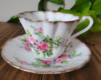 Puede incluir: Una taza de té y un platillo blancos con un patrón floral de rosas rosas y un borde dorado. La taza de té tiene un borde festoneado y el platillo tiene un borde festoneado.