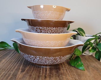 Può includere: Una pila di quattro ciotole Pyrex in varie tonalità di marrone e beige. Ogni ciotola presenta un motivo floreale bianco sul bordo. Le ciotole sono disposte su una superficie di legno, un set di utensili da cucina vintage.
