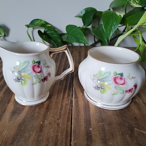 May include: A white cream jug and sugar bowl set with a floral design featuring pink roses, green leaves, and purple grapes. The set has a gold trim.