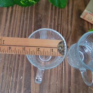 Puede incluir: Dos tazas de vidrio transparente con asas. Las tazas son texturizadas y tienen una superficie ligeramente ondulada. Una regla de madera se coloca sobre la parte superior de las tazas.