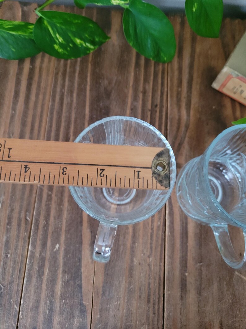 Puede incluir: Dos tazas de vidrio transparente con asas. Las tazas son texturizadas y tienen una superficie ligeramente ondulada. Una regla de madera se coloca sobre la parte superior de las tazas.
