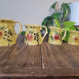 Peut inclure: Ensemble de quatre tasses à mesurer en céramique jaune avec un motif de fruits. Les tasses présentent un motif en relief de pommes, de raisins et de feuilles. Les tasses sont de tailles différentes, la plus grande tasse étant la plus haute.