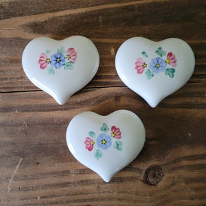 May include: Three white ceramic heart-shaped trinket boxes with painted floral designs. Each box features pink and blue flowers with green leaves.