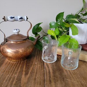 Puede incluir: Una tetera de cobre con un asa blanca y un dise&ntilde;o floral azul, dos tazas de vidrio transparente y una planta verde en maceta sobre una mesa de madera.