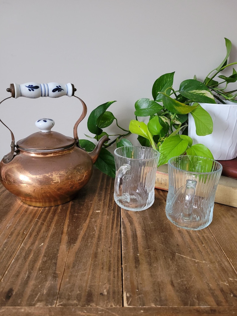 Puede incluir: Una tetera de cobre con un asa blanca y un dise&ntilde;o floral azul, dos tazas de vidrio transparente y una planta verde en maceta sobre una mesa de madera.