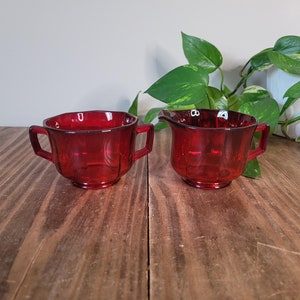 Puede incluir: Un juego de dos jarras de crema y azúcar de vidrio rojo. Las jarras tienen un diseño acanalado y están hechas de vidrio transparente con un tinte rojo.