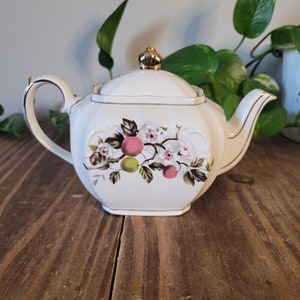 Könnte beinhalten: Eine weiße Teekanne mit goldenem Deckel und einem Blumendesign mit rosa und grünen Äpfeln und weißen Blüten. Die Teekanne hat einen quadratischen Korpus und einen goldenen Rand.