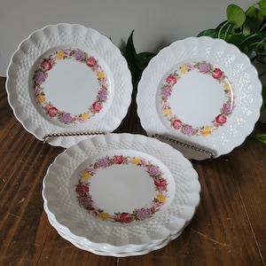 May include: Set of four white ceramic plates with a woven basket pattern and a floral border. The flowers are pink, purple, and yellow.