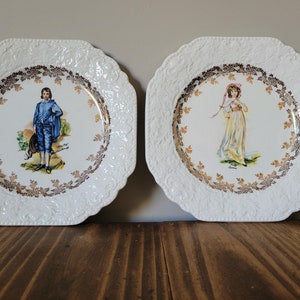 May include: Two white ceramic plates with gold trim and raised edges. One plate features a boy in blue clothing and the other features a girl in a white dress.