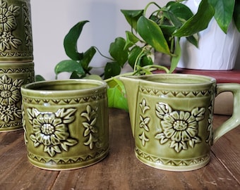 May include: A set of green ceramic tableware, including a creamer, a small cup, and stacked cups. Each piece features a raised floral design and a geometric border. The items are arranged on a wooden surface, with a plant in the background.