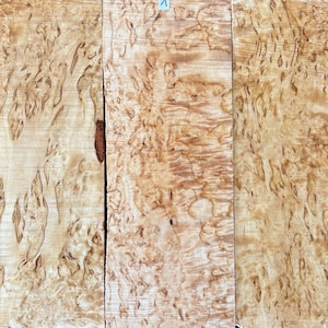 May include: Three wooden planks with a distinctive, swirling grain. The wood displays light tan and brown hues, with darker brown streaks and knots. One plank has a small label marked with the number "1".