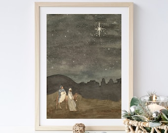 May include: Watercolor print of a religious scene with two figures on a donkey, under a starry night sky. The artwork is framed in a light wood frame. The scene depicts a landscape with a city silhouette and a bright star.
