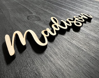 May include: A wooden sign with the name "Madison" written in a cursive font. The sign is painted black with the letters in a light wood color.