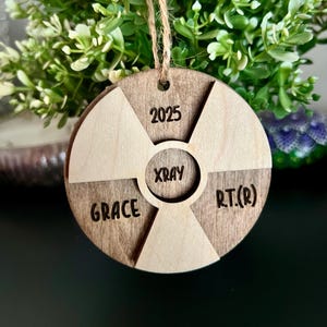May include: A wooden ornament shaped like a radiation symbol with the text "2025", "XRAY", "GRACE", and "RT.(R)" engraved on it.