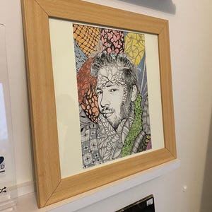 Könnte beinhalten: Ein gerahmtes Porträt-Kunstwerk, das eine Person mit Bart zeigt, umgeben von komplizierten, farbenfrohen Mustern in Orange, Gelb, Rosa und Grün. Das Bild befindet sich in einem hellen Holzrahmen mit einem weißen Passepartout-Rand.